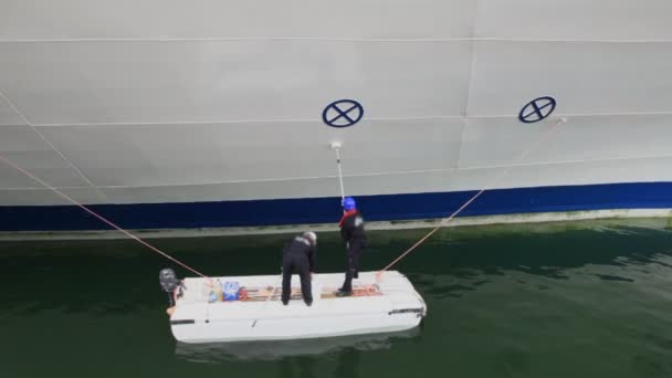 Twee werknemers staan op kleine boot en verf bestuur van enorme schip — Stockvideo