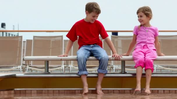 Two kids boy and little girl sit at pool edge near deckchairs — Stock Video