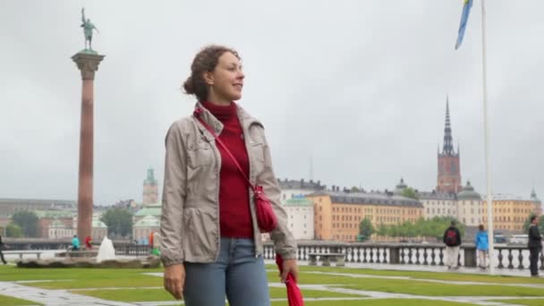 Женщина стоит на площади с фонтаном и памятником на речной набережной — стоковое видео