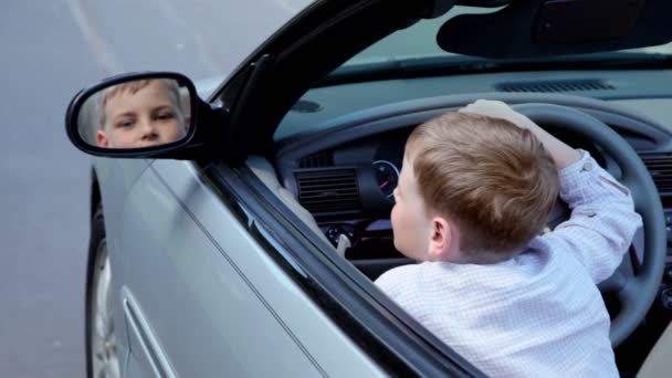 El niño se sienta en el coche en el lugar del conductor y mira en el espejo retrovisor — Vídeos de Stock