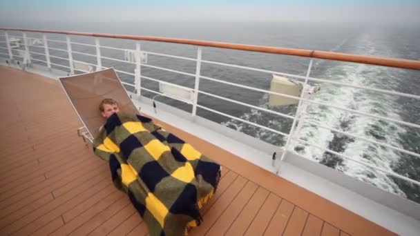 Little boy lies under plaid on deckchair at deck of ship — Stock Video