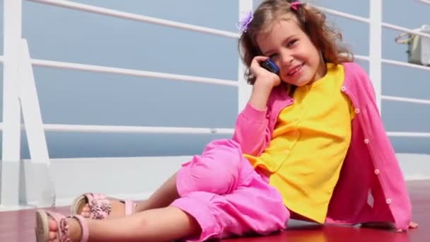 Little girl sits and talk by cell phone on deck near fence — Stock Video