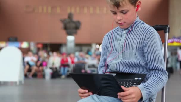 Boy hold netbook when sits on travelling bag at train station — Stock Video