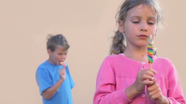 Küçük kız renkli şeker yiyor ve kardeşi arkasında aynısını yapıyor — Stok video