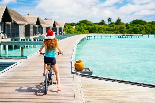 熱帯の島でサンタの帽子に乗って自転車に乗る若い女の子のバックビュークリスマス休暇を楽しんで — ストック写真