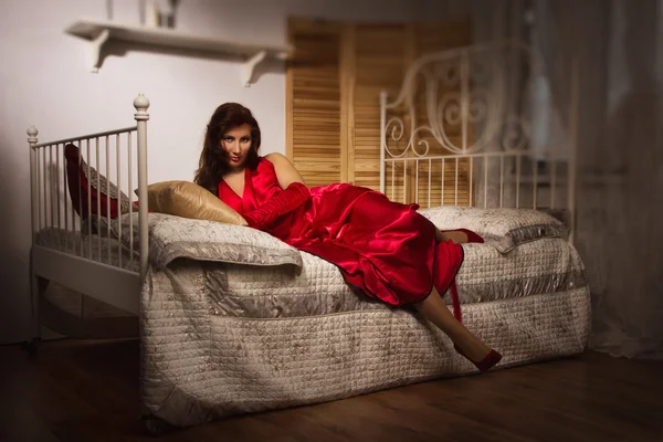 Sensuell brunett i en röd klänning som sitter på sängen — Stockfoto