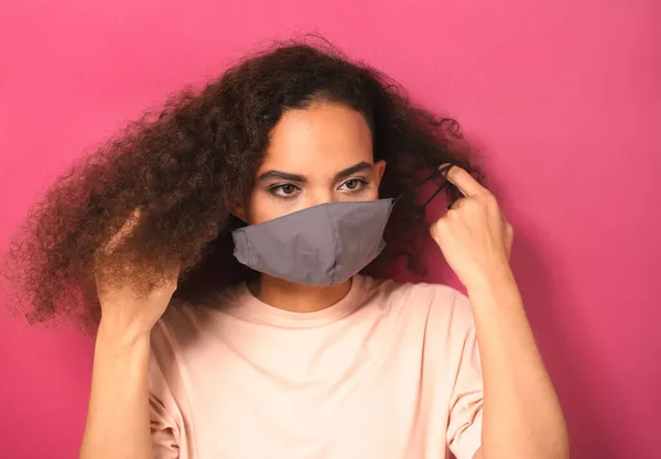 Colocando em máscara facial reutilizável afro-americano encantadora menina em t-shirt de pêssego, para evitar que os outros de corona COVID-19 e SARS cov 2 infecção isolada no fundo rosa — Fotografia de Stock