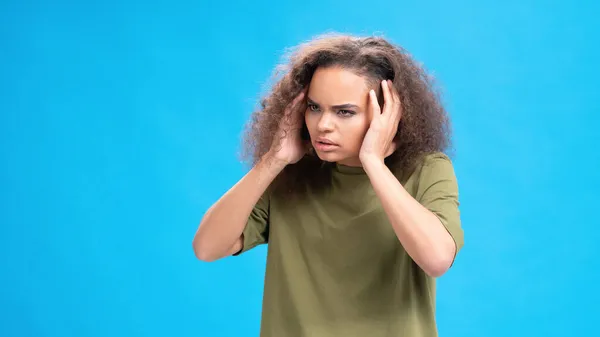 Ragazza afroamericana stressata o infelice che si tocca la testa soffrendo di mal di testa indossando una t-shirt olivastra isolata su sfondo blu. Il concetto di bellezza. Concetto sanitario — Foto Stock