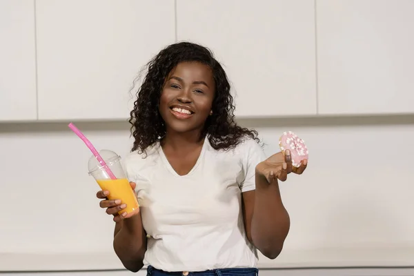 穿着白色T恤的非洲裔美国女人早餐喝橙汁配粉红甜甜圈。非洲女孩有一个橙汁玻璃杯和一个甜甜圈作为零食。特写。白人背景 — 图库照片