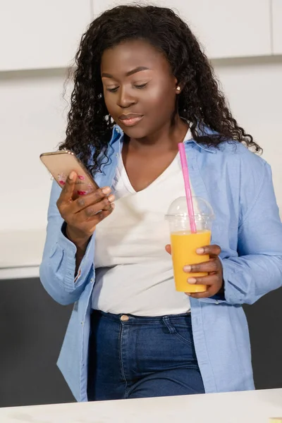 卷发的非洲女孩在她的智能手机上带着一杯橙汁在厨房里玩社交媒体。黑皮肤的女人喝新鲜的橙汁。四.后续行动. — 图库照片