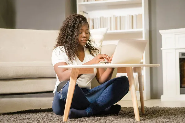 Όμορφη Αφρικάνα με διπλωμένα πόδια που κάθεται στο χαλί και δακτυλογραφεί στον υπολογιστή της. Η Αφρικάνα κοιτάζει μέσα από τη λίστα με τις ταινίες στο λάπτοπ της. Κοντινό πλάνο. Αρχική σελίδα Ιστορικό — Φωτογραφία Αρχείου