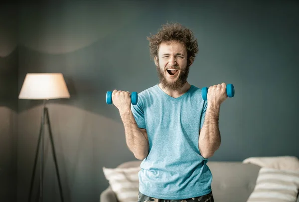 Funny Curly Guy disfruta bombeando sus bíceps por dos cascabeles especiales. El hombre divertido en camiseta azul tiene deporte en casa. Primer plano. —  Fotos de Stock