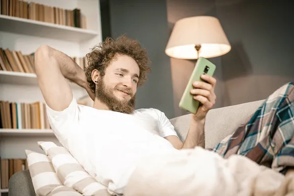 Ο σγουρομάλλης ξαπλωμένος στον καναπέ, κοιτάζοντας την οθόνη του τηλεφώνου και ελέγχοντας τα μέσα κοινωνικής δικτύωσης. Ρομαντικός τύπος που ψάχνει κοπέλα στο ίντερνετ. Κοντινό πλάνο — Φωτογραφία Αρχείου