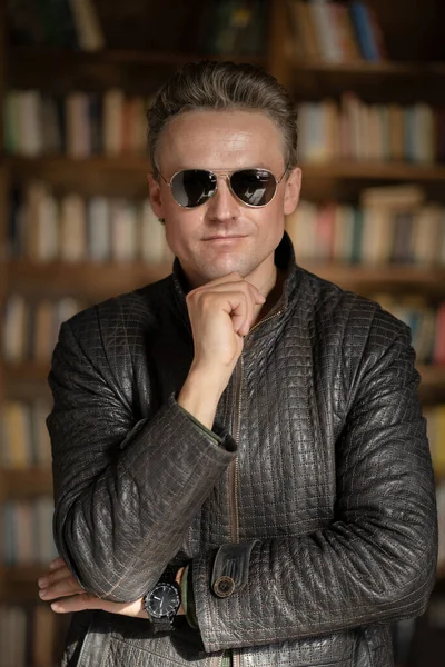 Brutal Fair-haired Guy in Modern Sunglasses Posing in his Business Office .White Man in Lather Jacket. Close-up Portrait. Book Shelves Background — Stock Photo, Image