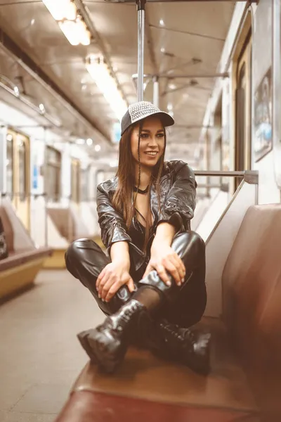 Улыбающаяся красивая брюнетка в современной латексной одежде сидит со сложенными ногами на сиденье в пустом вагоне метро. Милая женщина в подполье. Портрет крупного плана. Тонированная картинка — стоковое фото