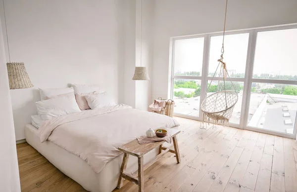 Quarto minimalista branco grande com cama de casal, banco de madeira, grandes molduras de janela, lâmpadas de teto de vime, cadeira de suspensão bege e piso de madeira lacada — Fotografia de Stock