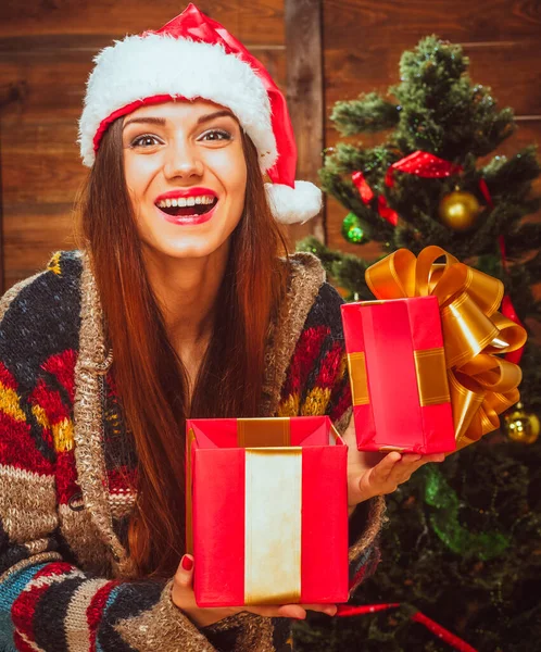 Caucasian Brunette Lady with a Wide Smile in a Casual Sweater and a Santa Hat Holds an Open Red Gift Box in her Hands Next to the Christmas Tree. Christmas Gifts, Christmas Mood. Wood Background — Stock Photo, Image