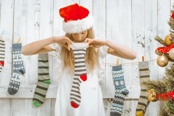 Vánoční punčocháče. Zvědavá dívka v bílých šatech a Santa Klobouk na hlavě se dívá do prázdné pruhované ponožky. Detailní záběr — Stock fotografie