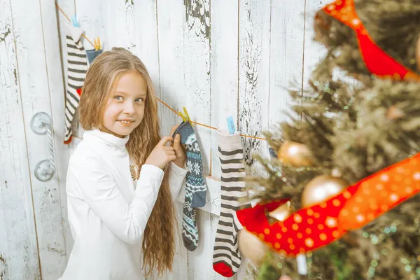 Linda chica caucásica en un puente blanco con largo pelo rubio cuelga los calcetines de Navidad. Medias de Navidad. Fondo de madera blanca. Primer plano — Foto de Stock