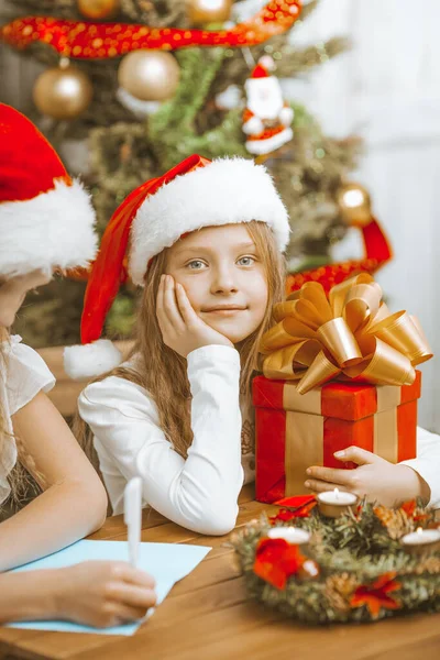 Little Sisters Girls in Christmas Hats Write a Letter to Santa Claus and Enjoy Christmas Eve. — Stock Photo, Image