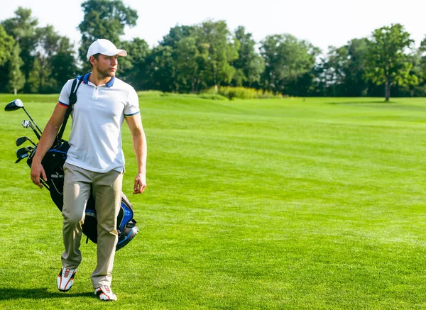 Молодий чоловік Голфер носить торбу з ключками для гольфу. Slim Man in White T-shirt, Golf Shoes, White Cap and Beige Pants is Going in the Green Turf Grass. Копіювальний простір. Повна довжина — стокове фото