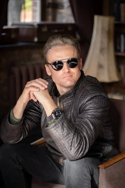 Sexy Blond Man in Stylish Sunglasses and a Leather Jacket Sits in an Armchair with Folded Hands. Портрет крупного плана. Винтажный интерьер — стоковое фото