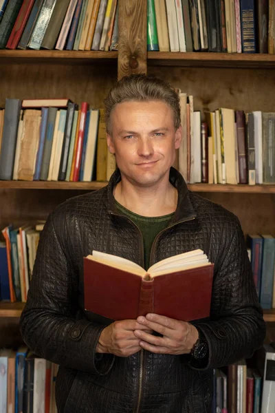 Un hombre guapo y seguro con un libro vintage rojo en sus manos se levanta contra el fondo de los estantes con libros de biblioteca privada. Primer plano Retrato — Foto de Stock