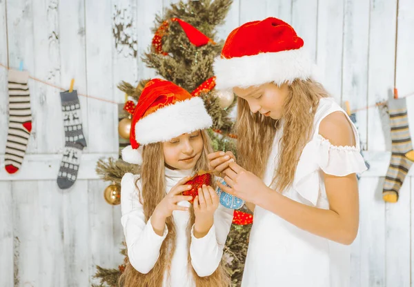 Teenage Girls in White Dresses and Red Hats Admire Christmas Decorations in their Hands. Preparing for Christmas, Christmas Eve. Close-up. Wooden Background — Stock Photo, Image