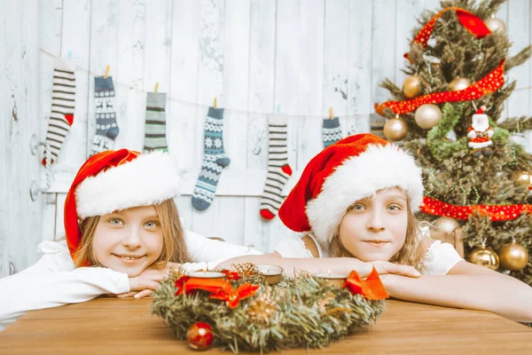 Two Cute Girls with Red Santa Hats are Sitting at the Table. Christmas Wreath on the Table. Room is Decorated for Christmas. Decorated Christmas tree. Christmas stockings on the Wall. Close-up — Stock Photo, Image