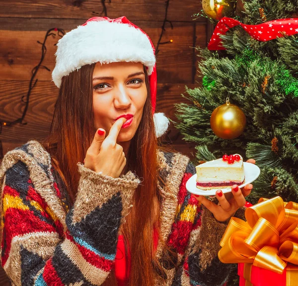Fényes hölgy kezében egy tányér egy szelet tortát, és kóstolja meg, ujj a szájban, Szenteste, karácsonyi vacsora, karácsonyi torta, karácsonyfa, karácsonyi ajándékok, fa háttér. Közelkép — Stock Fotó