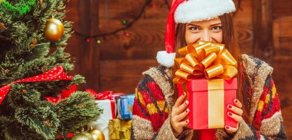 La dama de Navidad tiene una caja de regalo de la Plaza Roja. Mujer joven sostiene un regalo de Navidad en sus manos y cubre la parte inferior de su rostro. Copiar espacio. Fondo del árbol de Navidad. Pared de madera — Foto de Stock