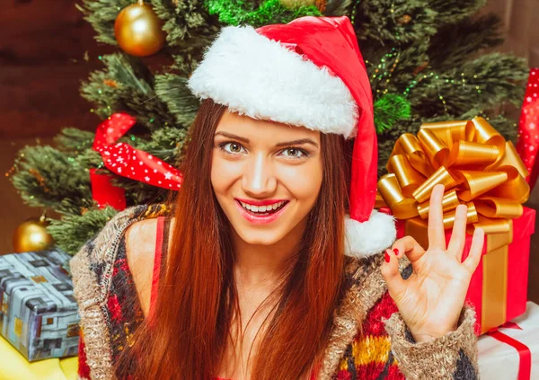 New Year Woman with Long Hair in a Red Hat Gazes to the Camera and Shows Hand Ok Εγγραφείτε. Πράσινο Διακοσμημένο Δέντρο της Πρωτοχρονιάς με πολλά κουτιά δώρων φόντο. Κοντινό πλάνο — Φωτογραφία Αρχείου