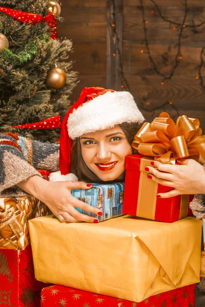 Υπέροχη νεαρή γυναίκα σε ένα κόκκινο Σάντα καπέλο κρατώντας μια δωδεκάδα των Χριστουγέννων πολύχρωμα δώρα. Διακοσμημένο χριστουγεννιάτικο δέντρο, ηλεκτρικό Γκάρλαντ στον τοίχο, φώτα. Κοντινό πλάνο. Πλαίσιο ξύλου — Φωτογραφία Αρχείου