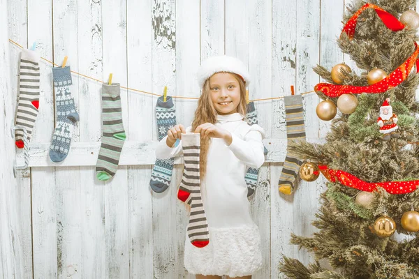 Beautiful Caucasian Girl of 9s in a Delicate White Dress and a Winter Hat Holds a Striped Christmas Sock for Gifts next to a Decorated Green Christmas Tree. Christmas Stocking . — Stock Photo, Image