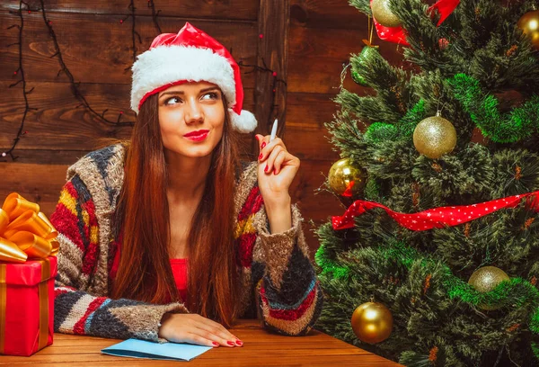 Очаровательная женщина в красной шляпе с белым мехом сидит за столом рядом с украшенной елкой и думает, что написать Санте. Крупный план — стоковое фото