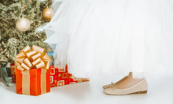 Girlish Legs in Beige Shoes and a Long White Tutu Skirt Stand on Floor Near Christmas Tree. Existuje mnoho dárků pod jedlí, čtvercové červené dárkové krabice se zlatou stuhou a Big Loopy luk. Detailní záběr — Stock fotografie