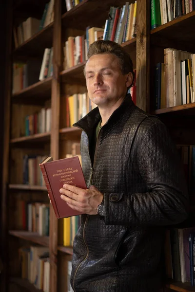 Zarif Özelliklere sahip yakışıklı aristokrat elinde eski bir kırmızı kitapla bir kitaplığın yanında duruyor, gözleri zevkten kapanıyor. Kütüphane Arkaplanı. Yakın plan Portre — Stok fotoğraf