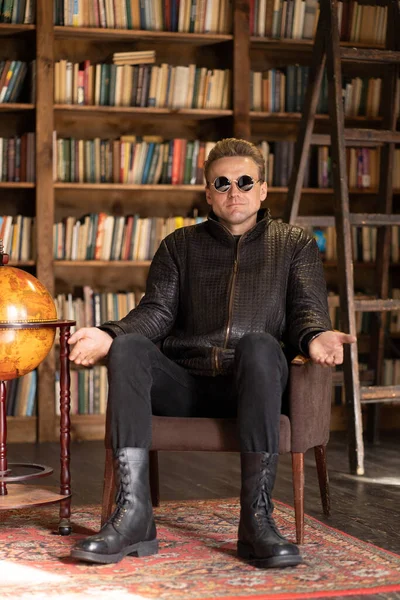 Man i hipster läderjacka och snygga solglasögon sitter i en retrofåtölj med öppna händer i Loft Studio. Bokhyllor Bakgrund. Närbild — Stockfoto