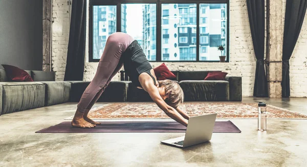 Nice Woman Practicing Yoga, Doing Down Dog Exercise, Watching Online Yoga Lessons by her Laptop, Yoga at Home. Close-up. Multi-storey Residential Building Background — Foto Stock