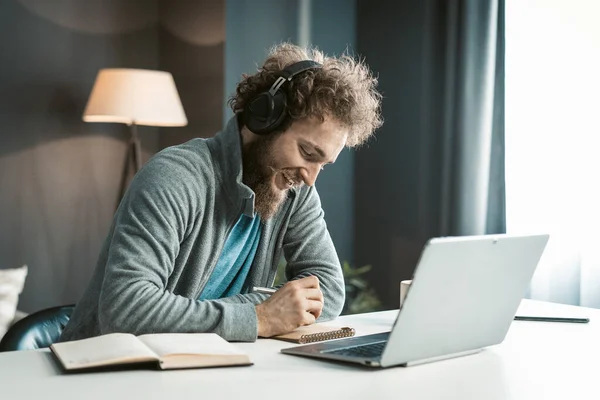 Young Student has an Online Video Lecture Uses a Laptop and Headphones, Takes Notes in a Notebook. Curly-Haired Guy sits in Earphones at the Table in his Office. Close-up — Stockfoto