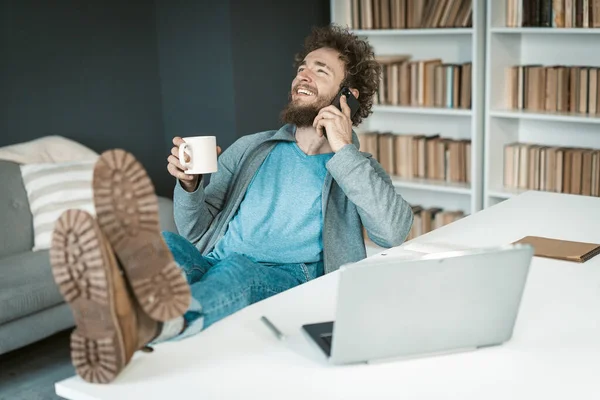 Empresario positivo en ropa casual tiene una conversación telefónica fácil con una taza de café en la mano y los pies sobre la mesa. Chico caucásico lindo que habla en Smartphone en la oficina. Primer plano — Foto de Stock