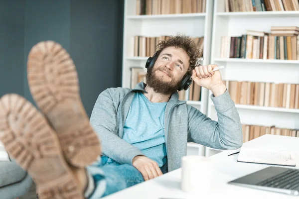 Lässiger Typ hört entspannende Musik über Kopfhörer in seinem Büro, Füße auf dem Tisch, Sohlen in die Kamera. Bücherregale im Hintergrund. Nahaufnahme — Stockfoto