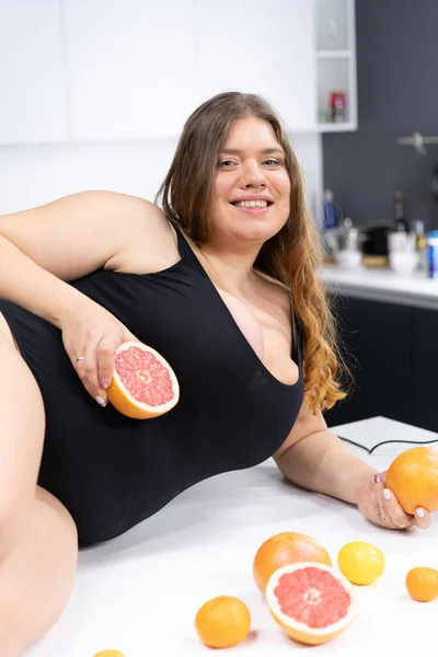 Weibliches Modell Plus Size in schwarzem Badeanzug posiert auf dem Küchentisch mit Zitrusfrüchten Orange, Grapefruit, Mandarine. Gesunde Ernährung, Körper positiv. Nahaufnahme — Stockfoto