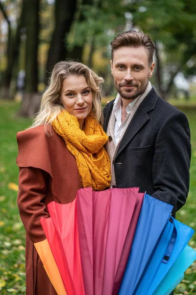 Hermosa pareja enamorada se para en el parque sosteniendo un paraguas de arco iris mirando a la cámara, Pareja familiar parque de otoño en tiempo lluvioso — Foto de Stock