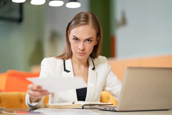 Mujer de negocios en una chaqueta blanca elegante se sienta en su escritorio con un ordenador portátil, miradas a la cámara y da papeles de negocios. Modelo Rubia Mujer con Ojos Azules Posando en una Chaqueta de Negocios en la Oficina. —  Fotos de Stock