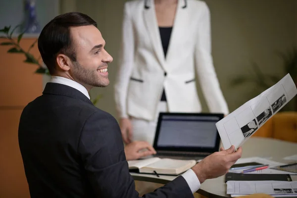 Empresário em um terno clássico e camisa branca senta-se em sua mesa em perfil para câmera. Chefe das Tarefas dos Delegados da Empresa no seu Gabinete. Trabalhador de colarinho branco. No fundo é uma senhora de negócios. — Fotografia de Stock