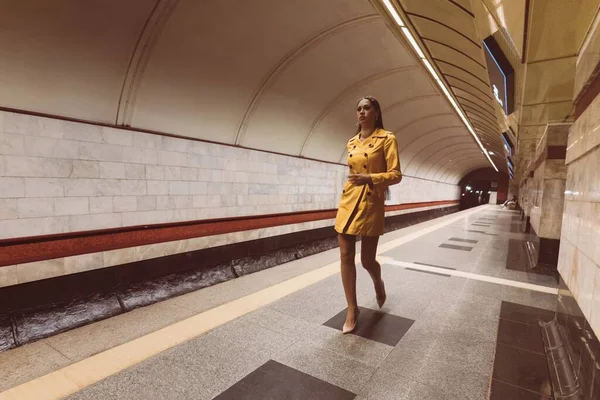 Marcher sur la plate-forme du métro belle jeune fille avec de longues jambes dans un manteau de printemps jaune et un sac à main blanc dans sa main. — Photo