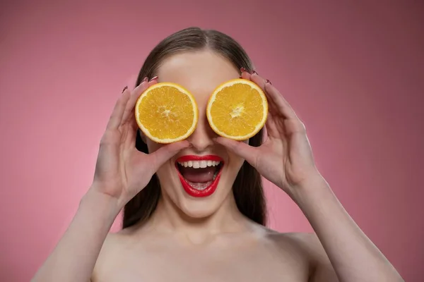 Engraçado bela beleza modelo jovem mulher segurando duas fatias de laranja suculentas como seus olhos ou óculos. Encantadora senhora engraçada alegre com lábios vermelhos e cabelos longos isolados no fundo rosa — Fotografia de Stock