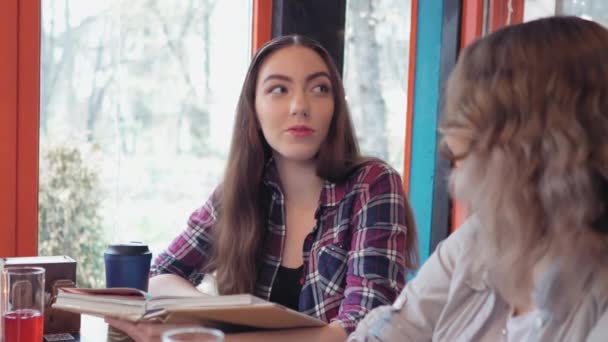 Zwei Studentinnen mit besten Freundinnen unterhalten sich in einem Café und teilen ihre Notizen, während im Hintergrund Lichter laufen, während offene Bücher und Laptop auf dem Tisch liegen. 4K-Filmmaterial Lizenzfreies Stock-Filmmaterial