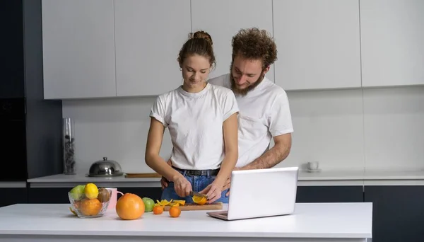 Ευτυχισμένη οικογένεια στο νέο τους σπίτι. Όμορφος άντρας αγκαλιάζει τη γυναίκα του χαμογελώντας κοιτάζοντας την κάμερα. Όμορφο νεαρό ζευγάρι μιλάει σε βιντεοκλήση χρησιμοποιώντας το laptop. Νεαρό ζευγάρι μαγείρεμα υγιεινό φαγητό στην κουζίνα στο σπίτι — Φωτογραφία Αρχείου